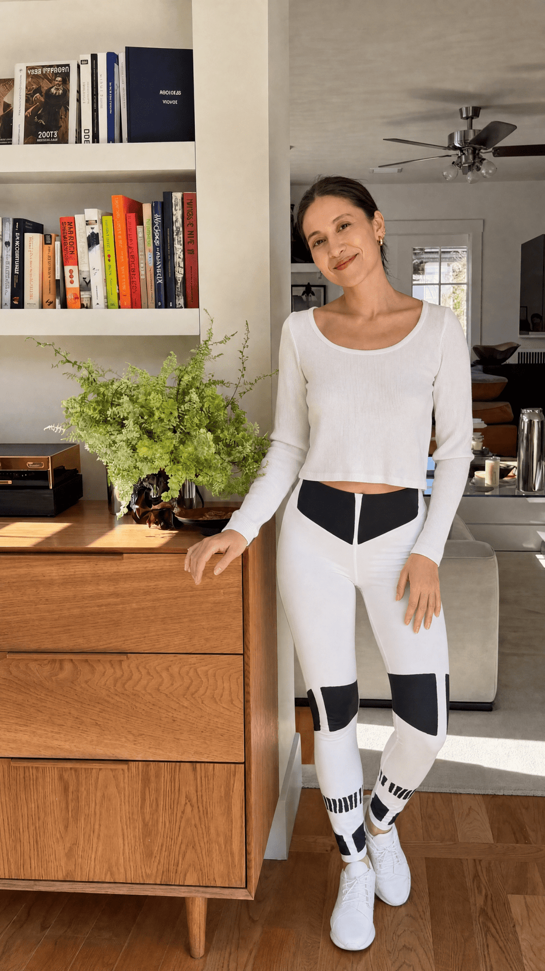 Woman in a white outfit standing in a room with a wooden dresser and books.