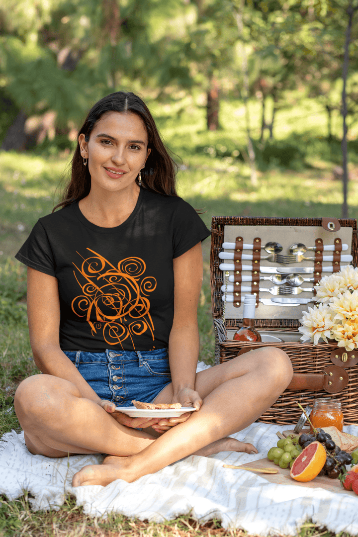 A woman sits outdoors on a blanket with a plate of food, wearing the Trend - Abstract T-Shirt by Miamiabstract.com and denim shorts. Beside her is a picnic basket filled with fruit, utensils, and flowers.