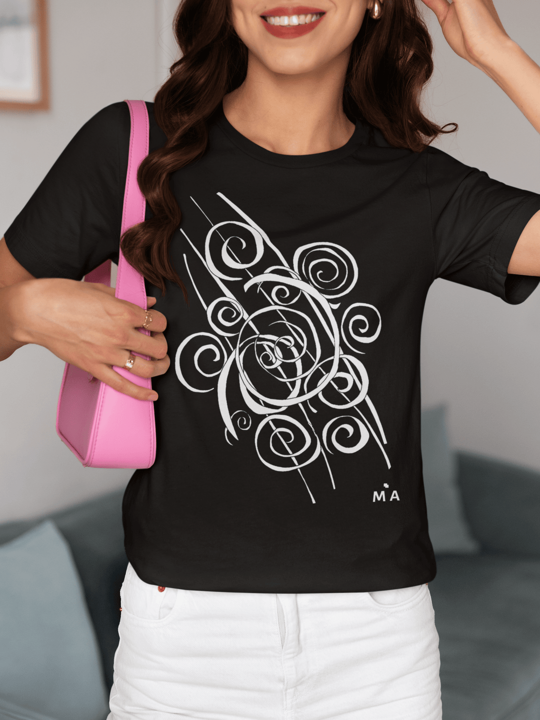 A smiling woman with wavy brown hair wears the Trend - Abstract T-Shirt from Miamiabstract.com, featuring a black design with a white swirl and “M A” initials. She holds a pink shoulder bag while sitting on a couch.