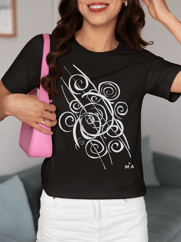 A smiling woman with wavy brown hair wears the Trend - Abstract T-Shirt from Miamiabstract.com, featuring a black design with a white swirl and “M A” initials. She holds a pink shoulder bag while sitting on a couch.