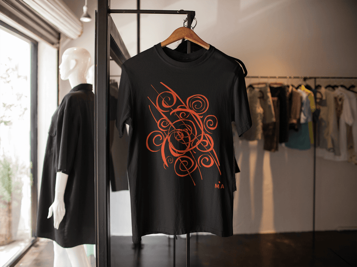The Trend - Abstract T-Shirt by Miamiabstract.com, featuring a bold black and orange swirl silk screen print, hangs on a wooden hanger in a boutique with abstract fashion pieces displayed in the background.