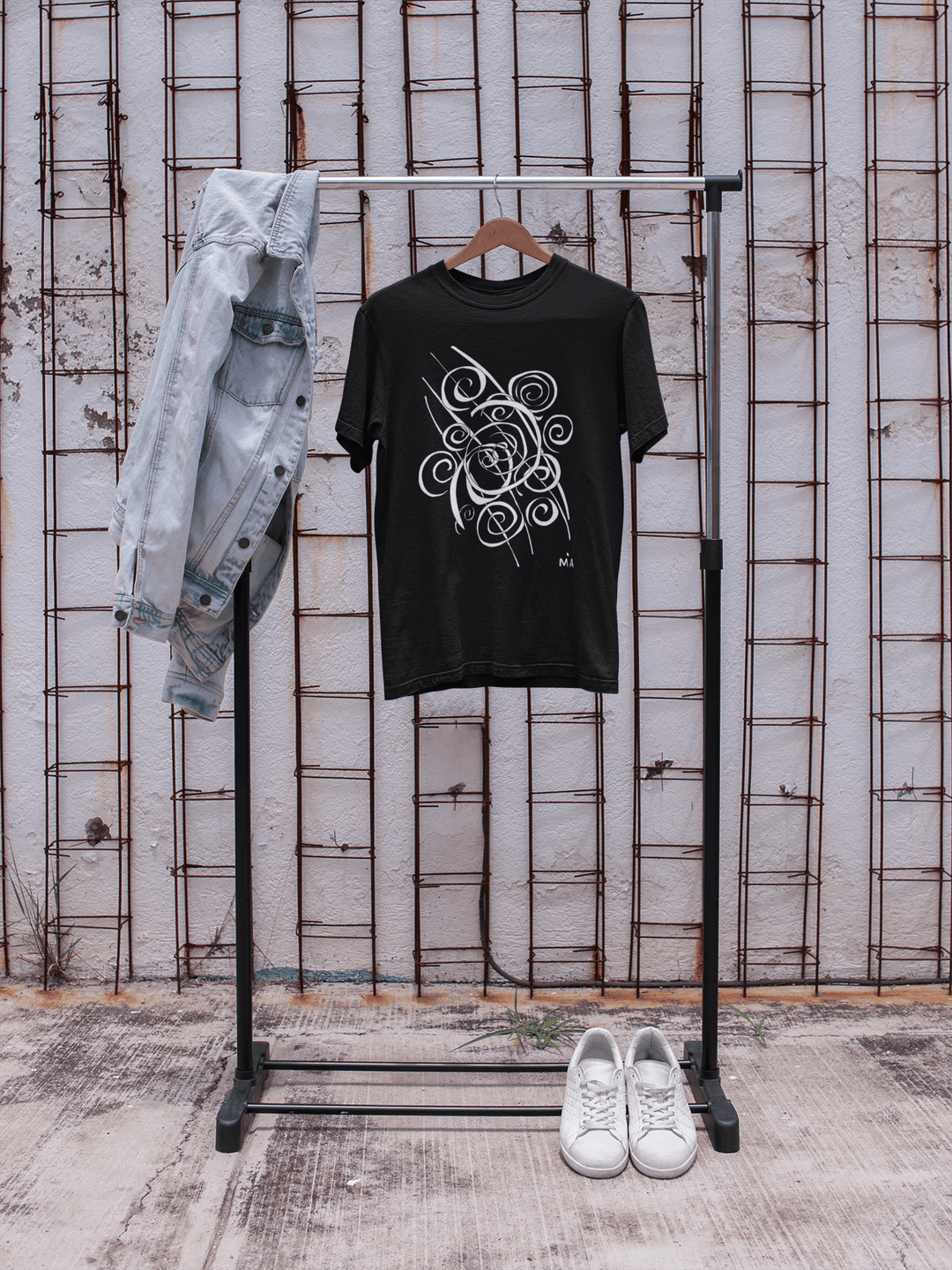 The Trend - Abstract T-Shirt by Miamiabstract.com, featuring a black silk fabric with a white swirl design, hangs next to a light blue denim jacket, while white sneakers rest below by a wall with exposed metal rods.