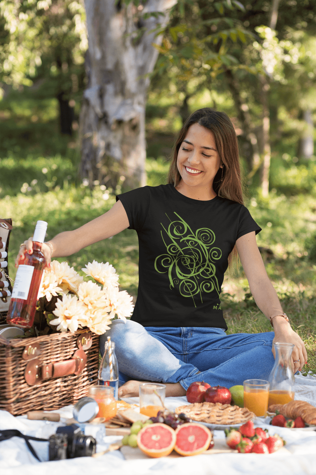 Wearing the Trend - Abstract T-Shirt by Miamiabstract.com, a woman enjoys a picnic on a blanket, smiling as she reaches for rosé wine near a wicker basket with flowers and fruits, surrounded by greenery and sunlight.